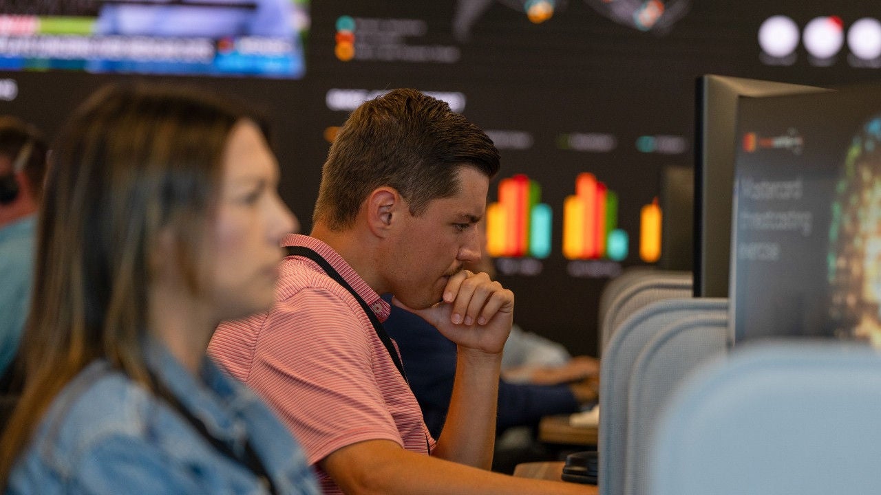 Two Mastercard employees look at their computers in front of a large screen displaying a bar graph.