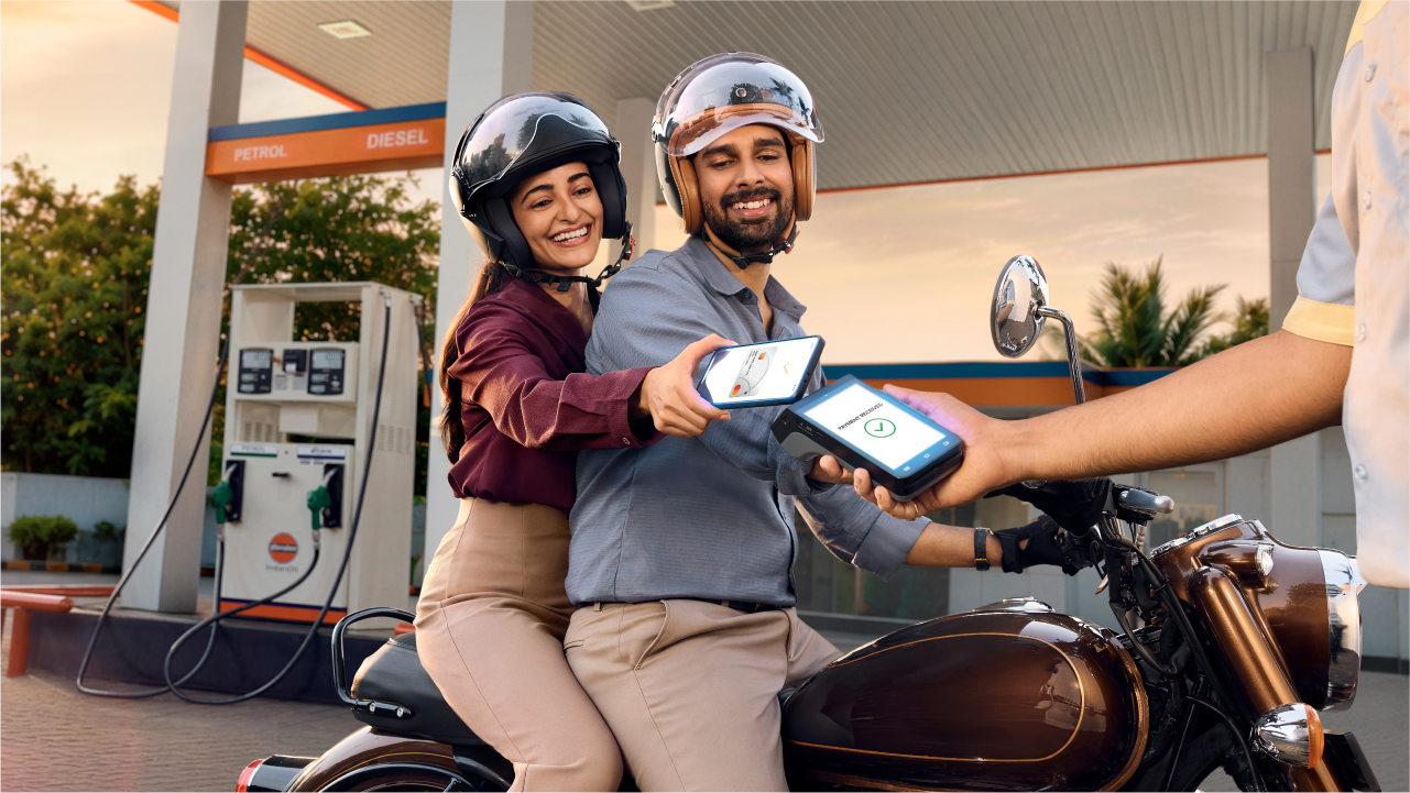 Indian couple sitting on a bike, at a petrol pump. Wife sitting at the back of the bike and making a payment by tapping her Mastercard on POS machine.