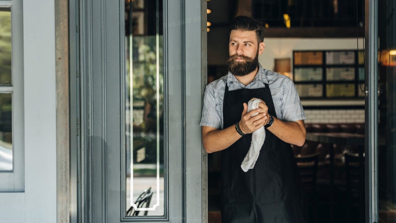 Kávézóban álló barista kötényben, kezében törlőkendővel, az ajtóban kifelé tekintve.