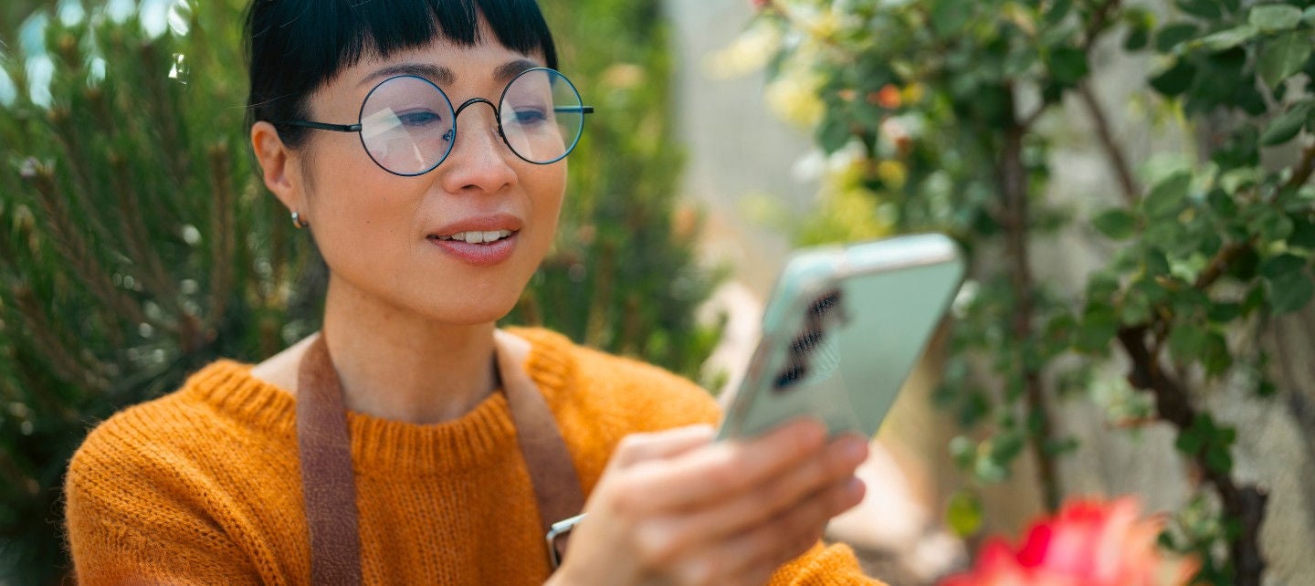 A woman in an apron holds a phone up to a flower. 