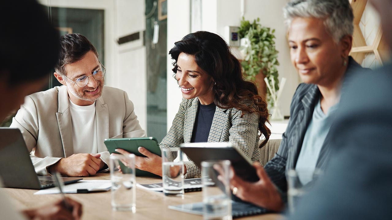 Four people in business-casual attire sitting around a table with tablets in a bright office, discussing a project.