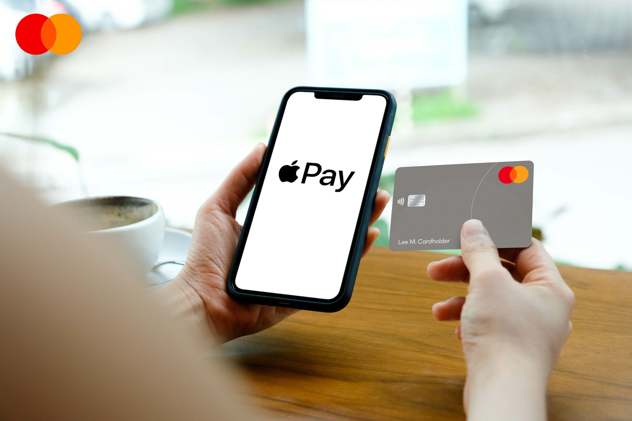 One hand holds a phone displaying a payment app screen while the other holds a grey payment card.