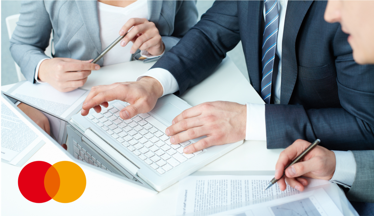 A close-up of three people in business attire at a desk, one typing on a laptop while the others hold pens over documents.