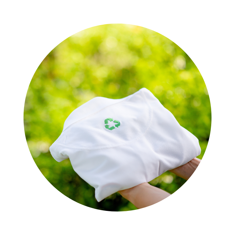 Kézben tartott fehér ruhadarab újrahasznosítási jellel, zöld, természetes háttér előtt, a fenntartható öltözködést szimbolizálva.