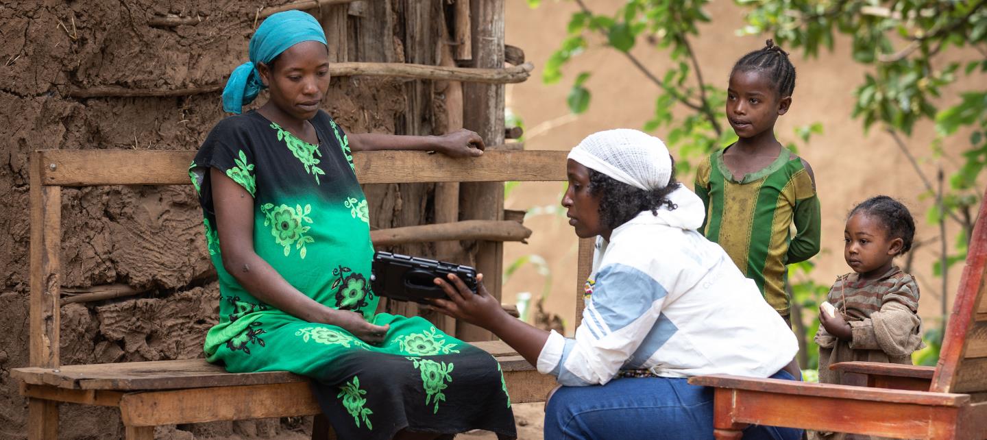 A community health workers in Ethiopia shows a tablet to a patient while two children look on. 