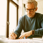 Man with glasses, writing diligently at a desk by a window.