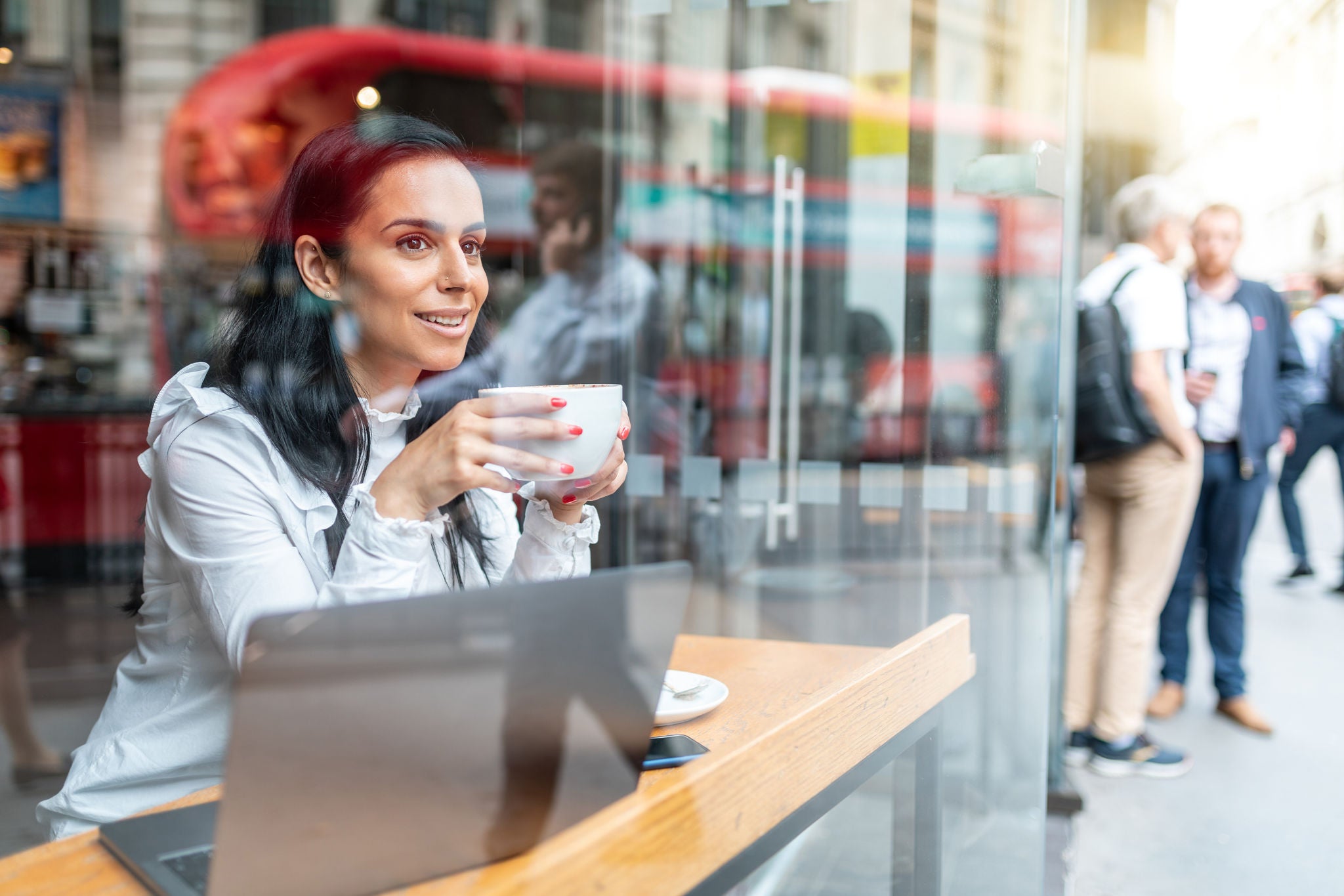 Professionelle Frau lächelt, trinkt Kaffee und arbeitet an einem Laptop am Fenster eines Cafés, mit Spiegelungen einer Stadtstraße und eines roten Doppeldeckerbusses.