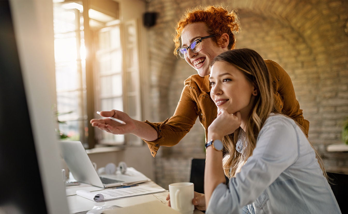 Two women sit at a desk, focused on a computer, engaged in a discussion or collaboration.