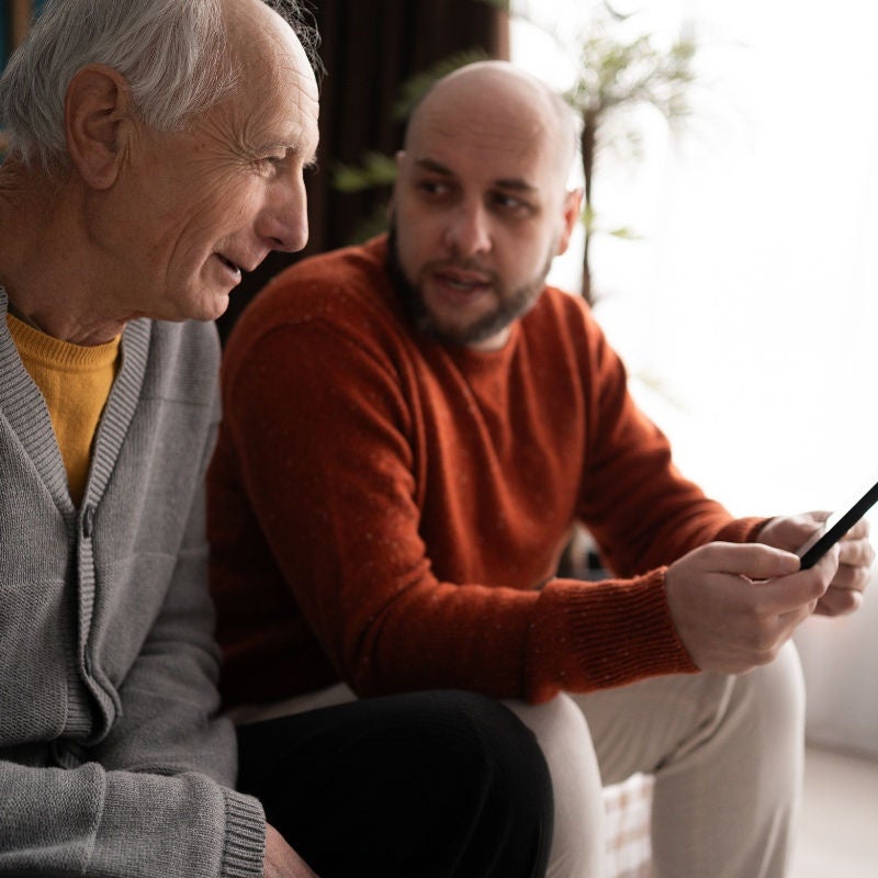A younger man holds a phone and talks to an older man on a couch. 