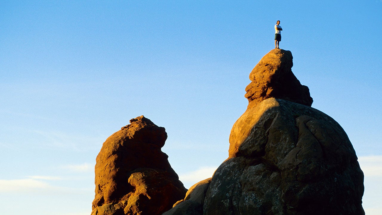 En person i silhuet står på toppen af en stor klippe mod en klar blå himmel.