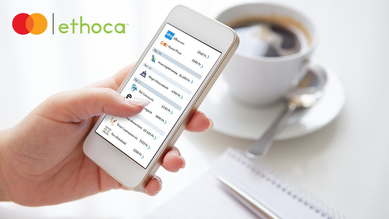 Egy kézben tartott okostelefonon az Ethoca alkalmazás tranzakciós listája látható, az asztalon kávéscsésze és jegyzetfüzet mellett.