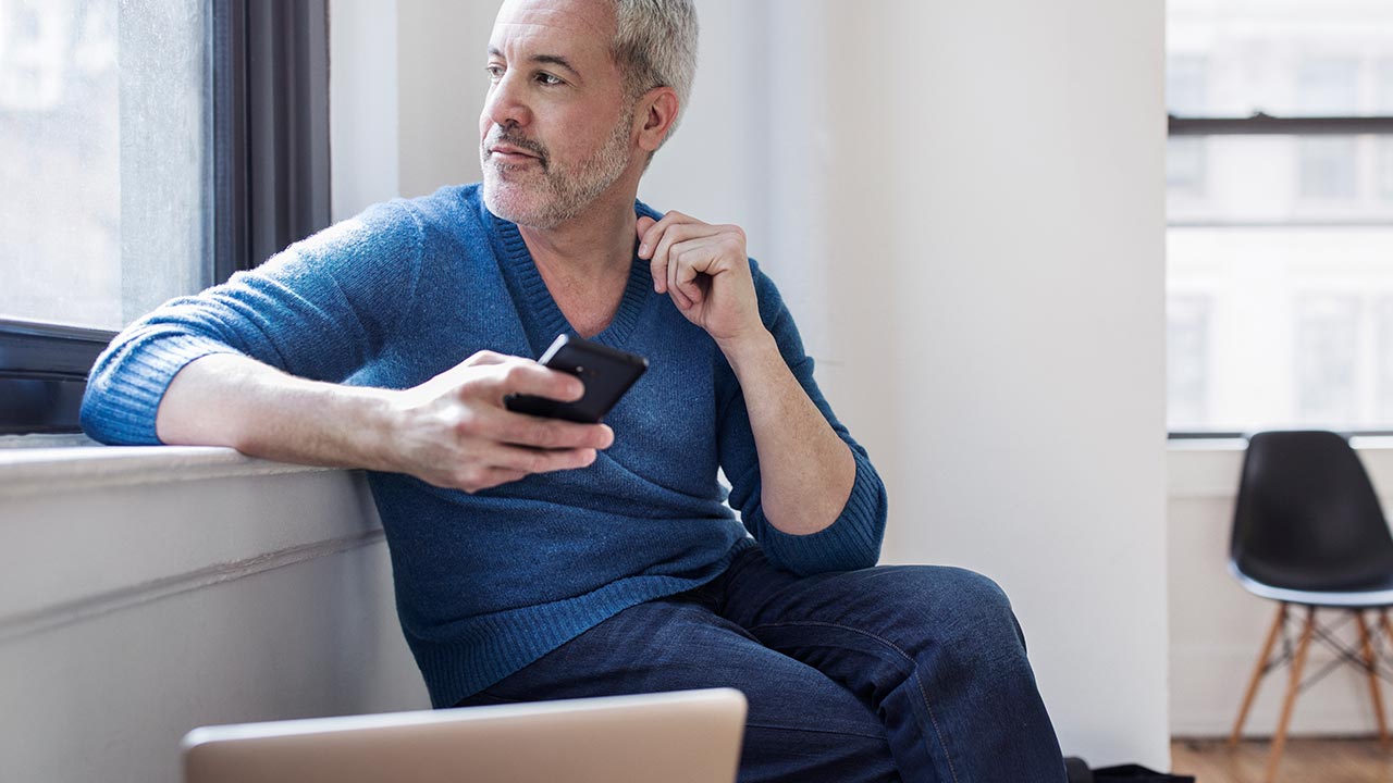 Középkorú férfi ül az ablakpárkányon, okostelefont tart a kezében, miközben kifelé néz; előtte egy nyitott laptop látható.