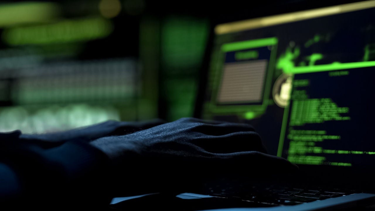 A person's hands hover over a keyboard on a laptop with coding on the screen in a darkened room. 