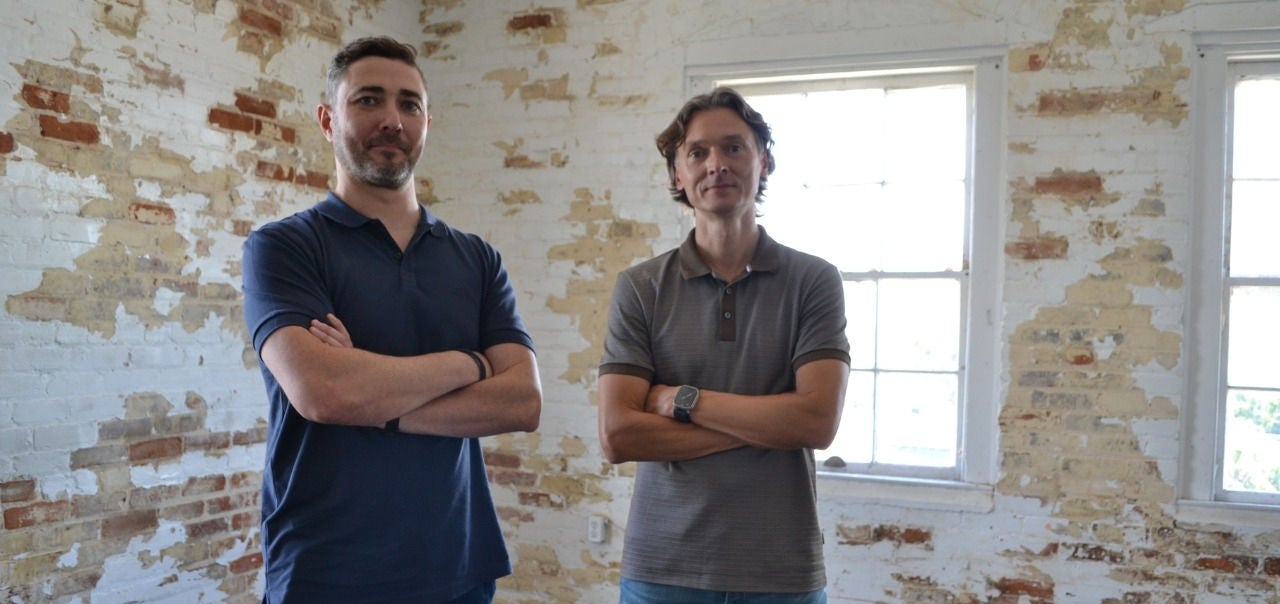 Ilya Volovik and Andrei Barysevich stand, arms crossed in a room with distressed paint on brick walls in front of a window. 
