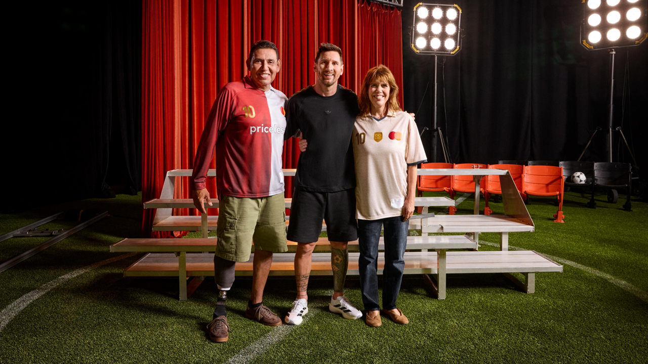 Lionel Messi smiling as he exchanges jerseys with a female fan. Messi holds a bright green, signed "Messi 10" MasterCard jersey, while the fan hands over a dark shirt on a stage with a red curtain backdrop.