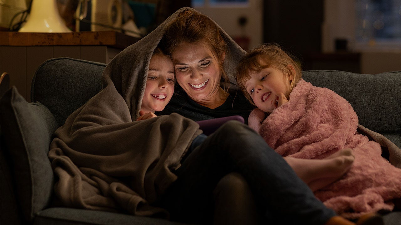 Family under a blanket, using tablet and sitting on sofa.