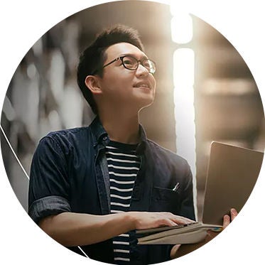 A man wearing glasses holds a laptop, looking focused and engaged in his work.