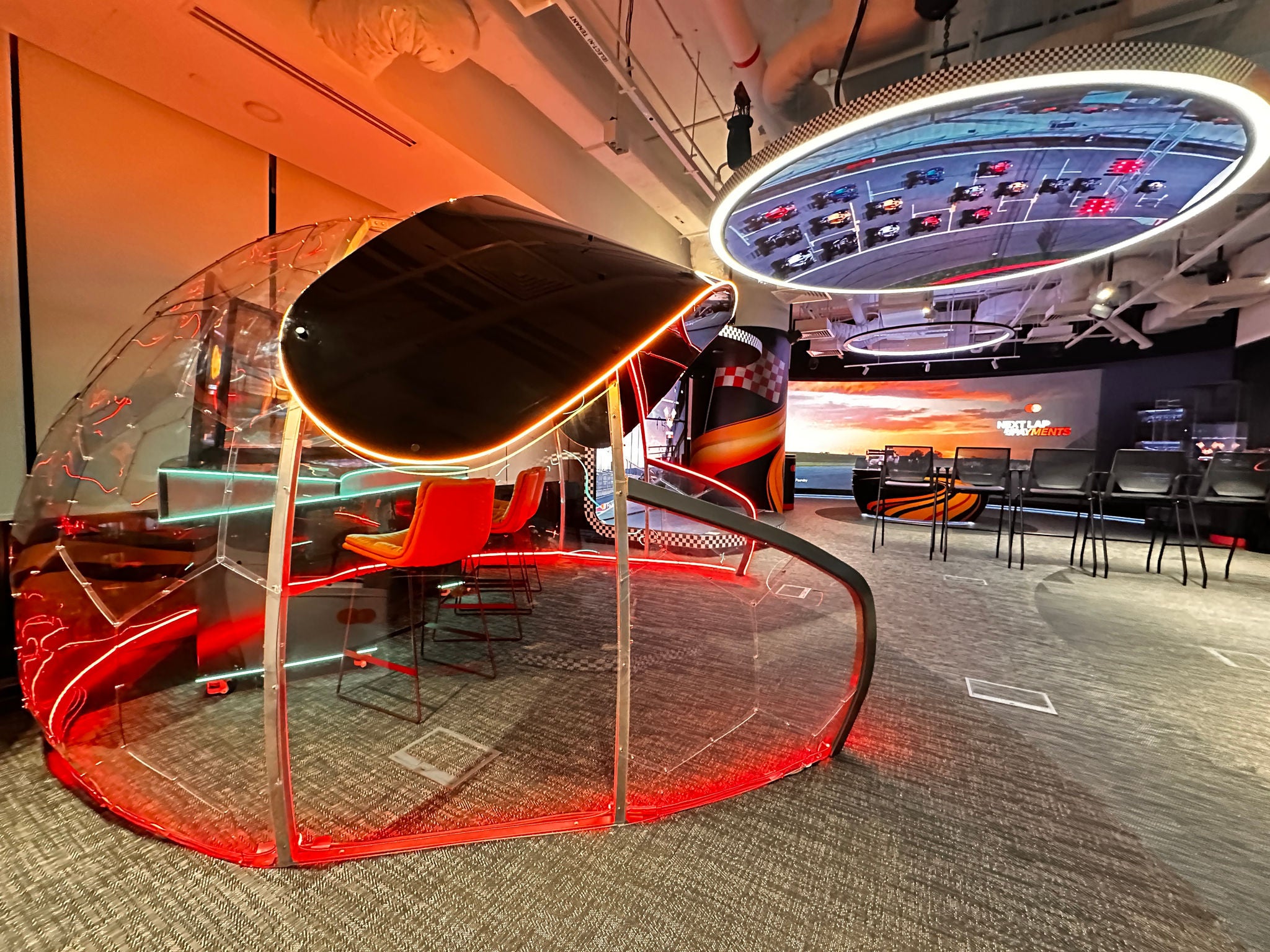 An interior exhibition space with a transparent dome seating pod, orange neon lighting, a circular ceiling screen showing race cars, and rows of chairs beside a large wall display.