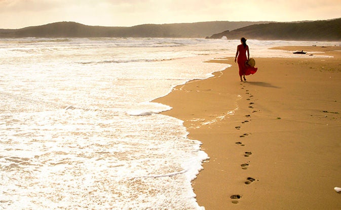 A woman walks along the beach, leaving footprints in the sand under a clear blue sky.