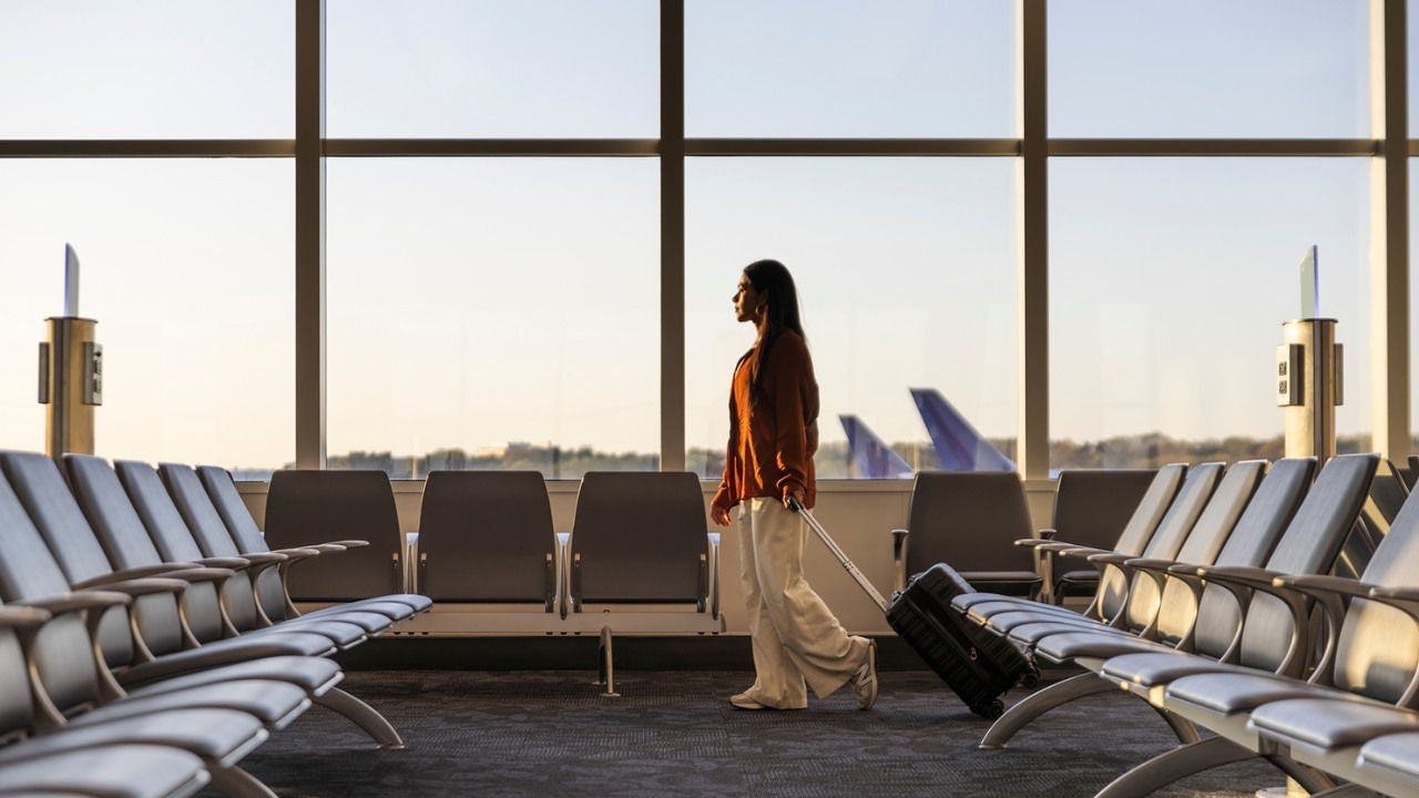 A woman pulls a suitcase through an airport where the fins of airplanes can be seen through the windows. 