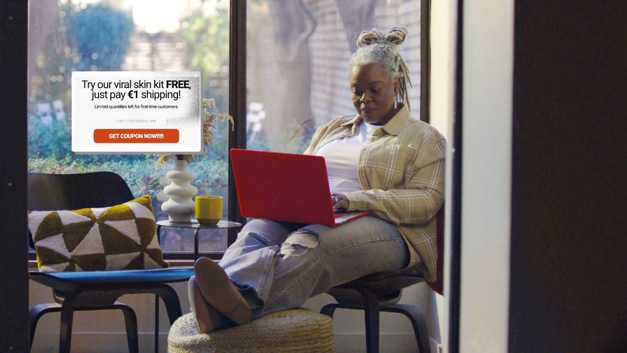 A woman seated on a chair with her feet on a hassock looks at a laptop with an overlay of a scam ad. 