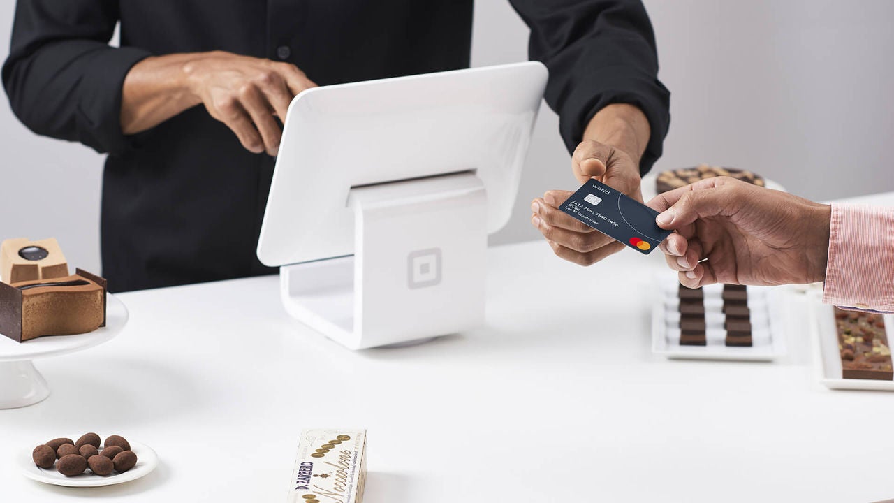 Человек передает кредитную карту кассиру у современного POS-терминала. Действие, по всей видимости, происходит в пекарне или кондитерской, на прилавке выставлены различные шоколадные конфеты и пирожные. Сцена запечатлела момент совершения транзакции, подчеркивая использование бесконтактной технологии оплаты. Оба человека частично видны, фокус на их руках и процессе оплаты.