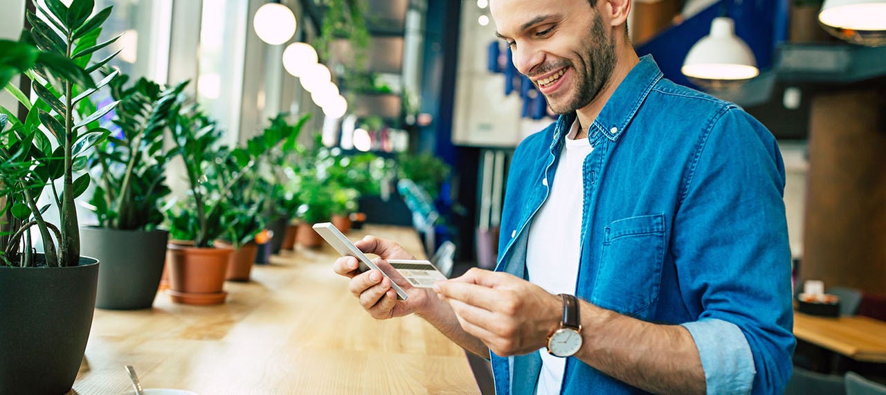 Man holding a credit card and smartphone while making an online payment at a café