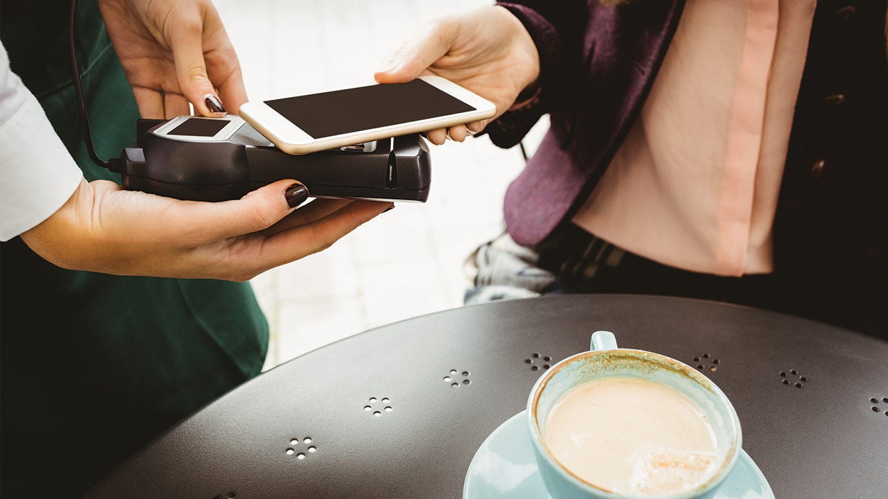 Kávézóban egy vendég okostelefonnal érintésmentesen fizet egy hordozható bankkártya-terminálnál, az asztalon egy csésze kávé látható.