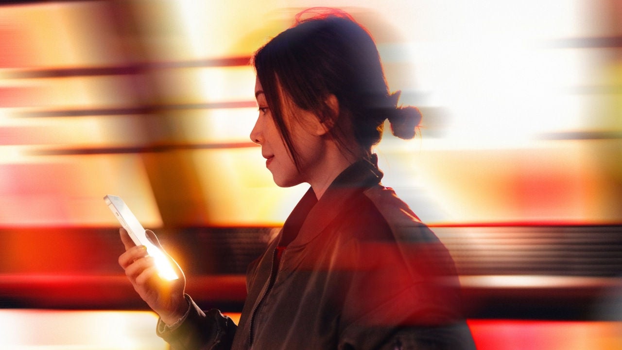 A woman walks while looking at a phone in front of a red and yellow background. 