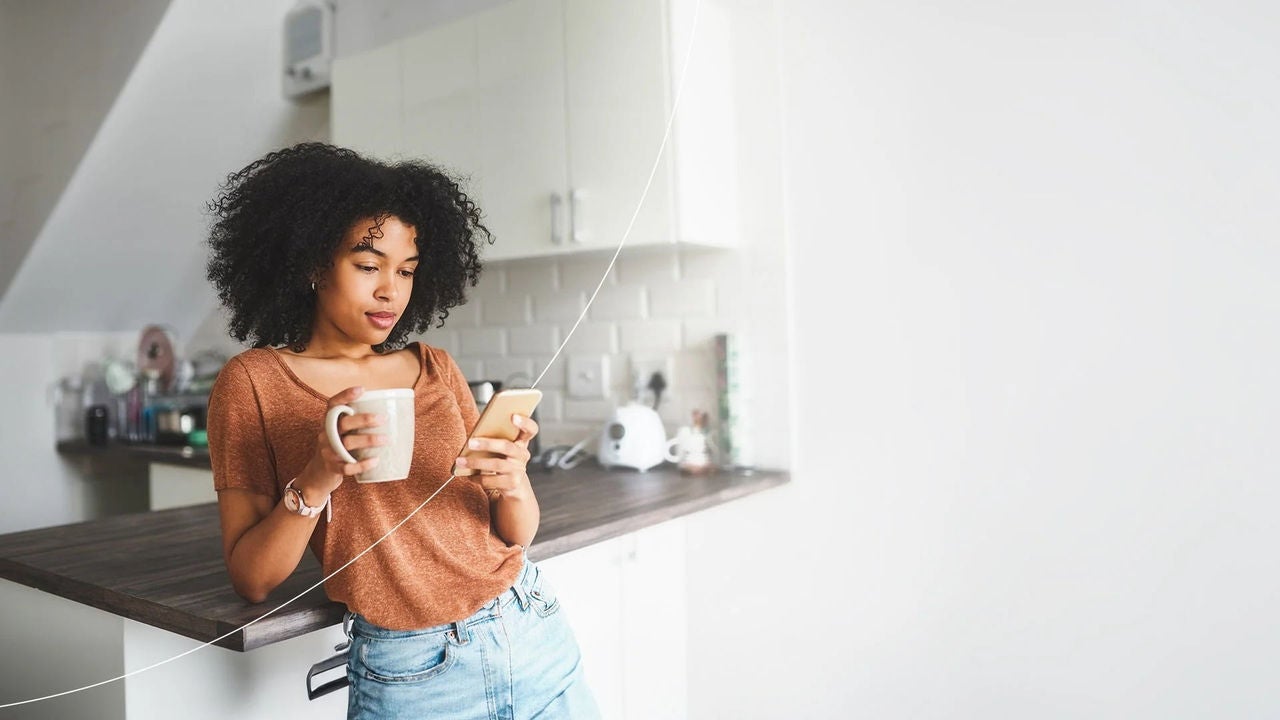 אישה עם כוס קפה משתמשת בטלפון במטבח.