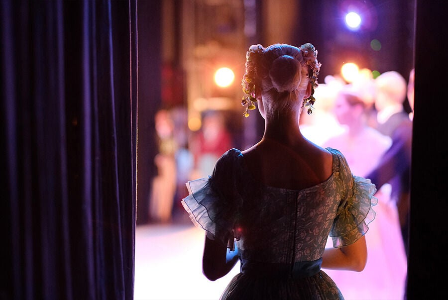 A ballet dancer in a light blue costume waits backstage, looking toward the softly lit stage where other performers are blurred in the background under warm, glowing lights.