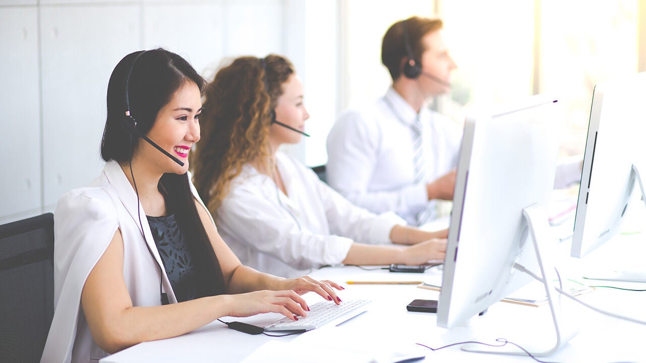 Ügyfélszolgálati munkatársak headsettel dolgoznak egy irodában; az előtérben egy mosolygó nő gépel a számítógépen.