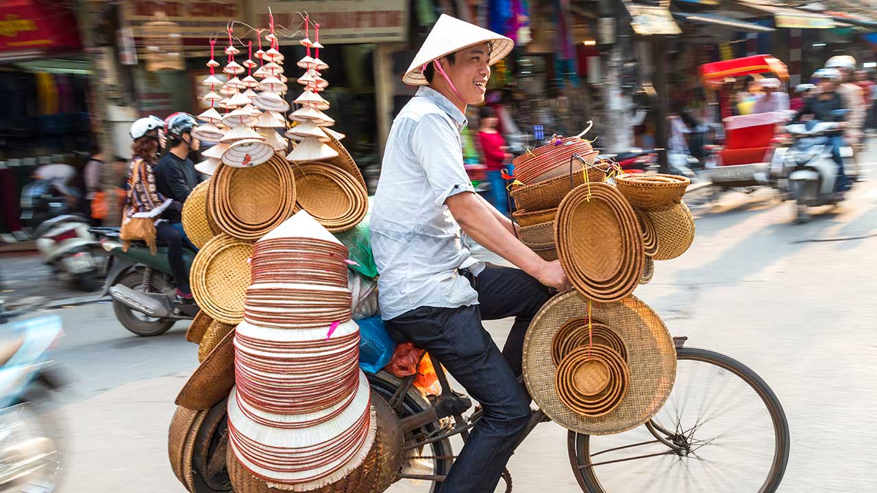 Mosolygó utcai árus kerékpáron haladva, fonott kosarakat és hagyományos kalapokat szállít egy forgalmas városi utcán.