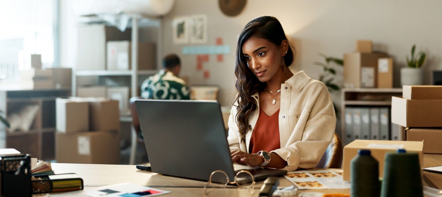 A woman business owner looks at her laptop in an office full of packages. 