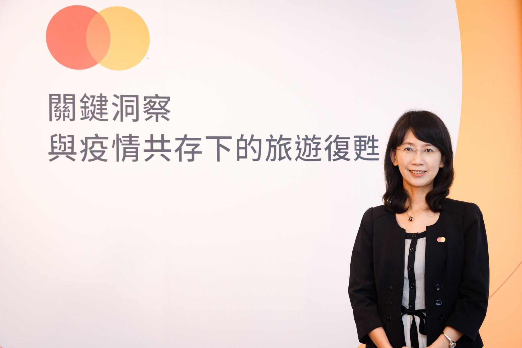 A woman in a black jacket stands smiling in front of a white and orange backdrop with Chinese text and a brand logo.
