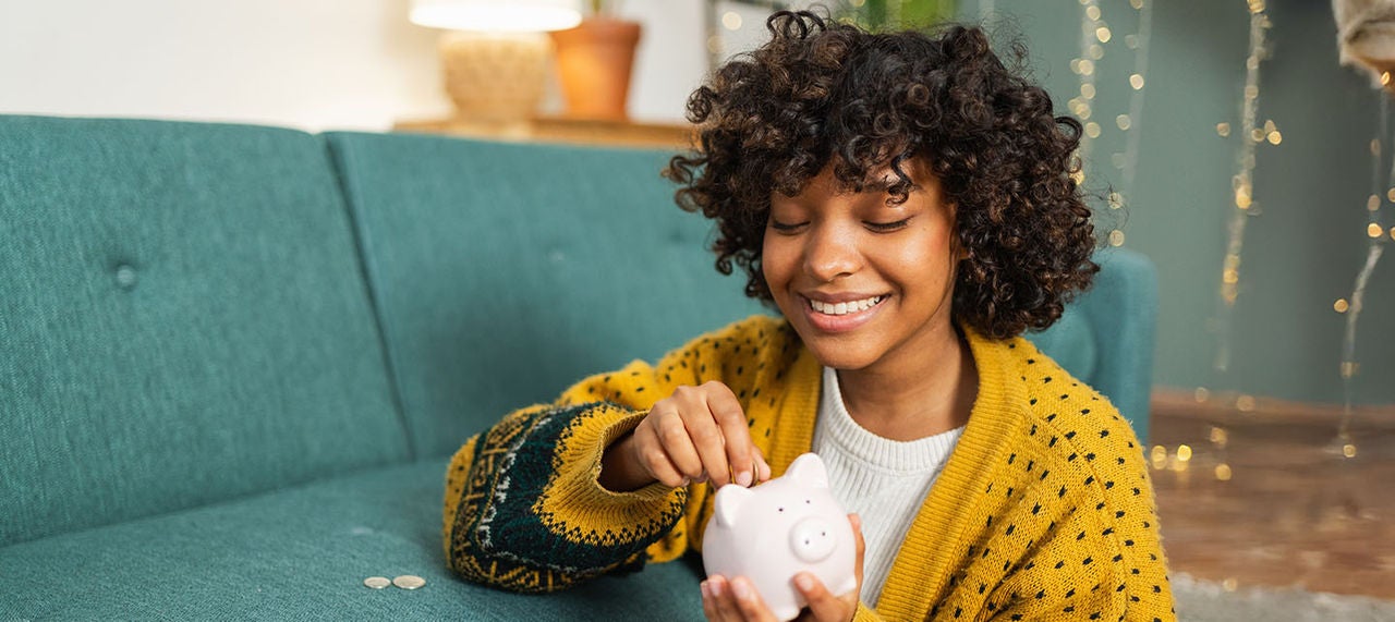 Happy young woman sitting on floor putting coins into a piggy bank.