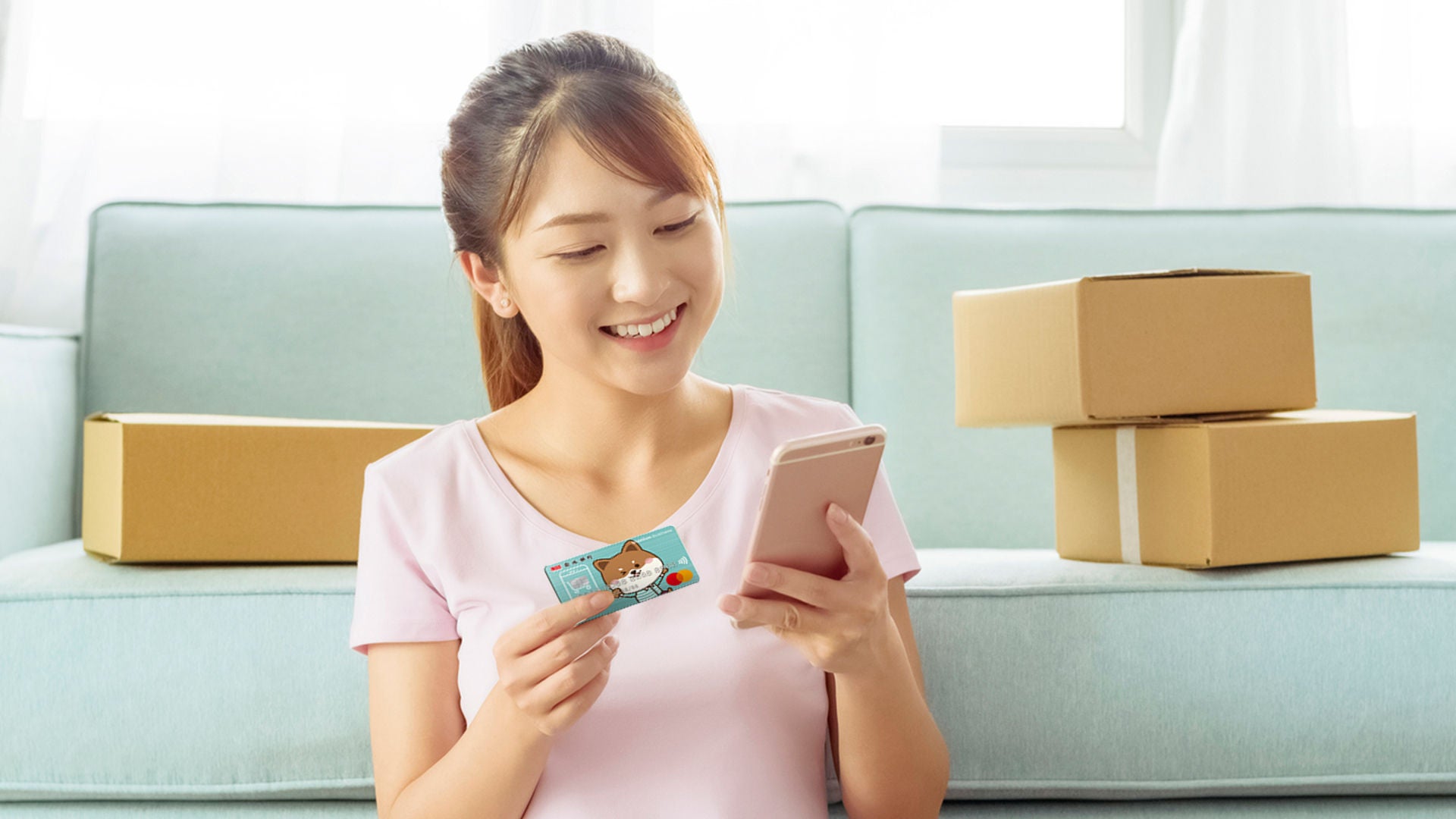 A young woman in a pink top sits in front of a sofa holding a payment card and a phone, with cardboard parcels beside her.