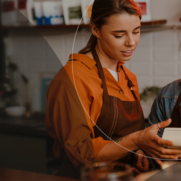 A woman wearing an orange hoodie and brown apron works behind a café counter, looking at a tablet while preparing an order.