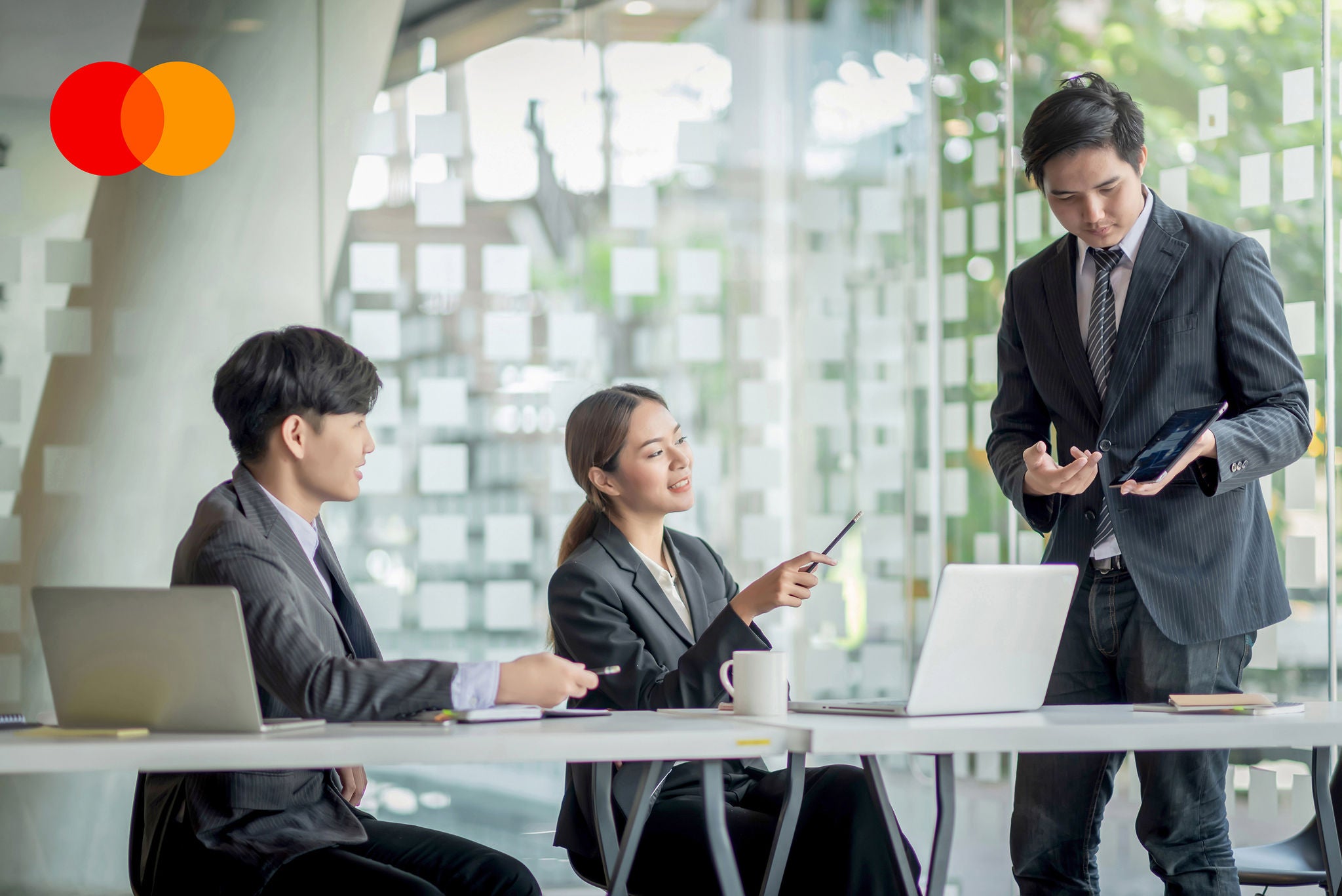 Three people in business suits discuss around a white table with laptops in a bright glass-walled office.