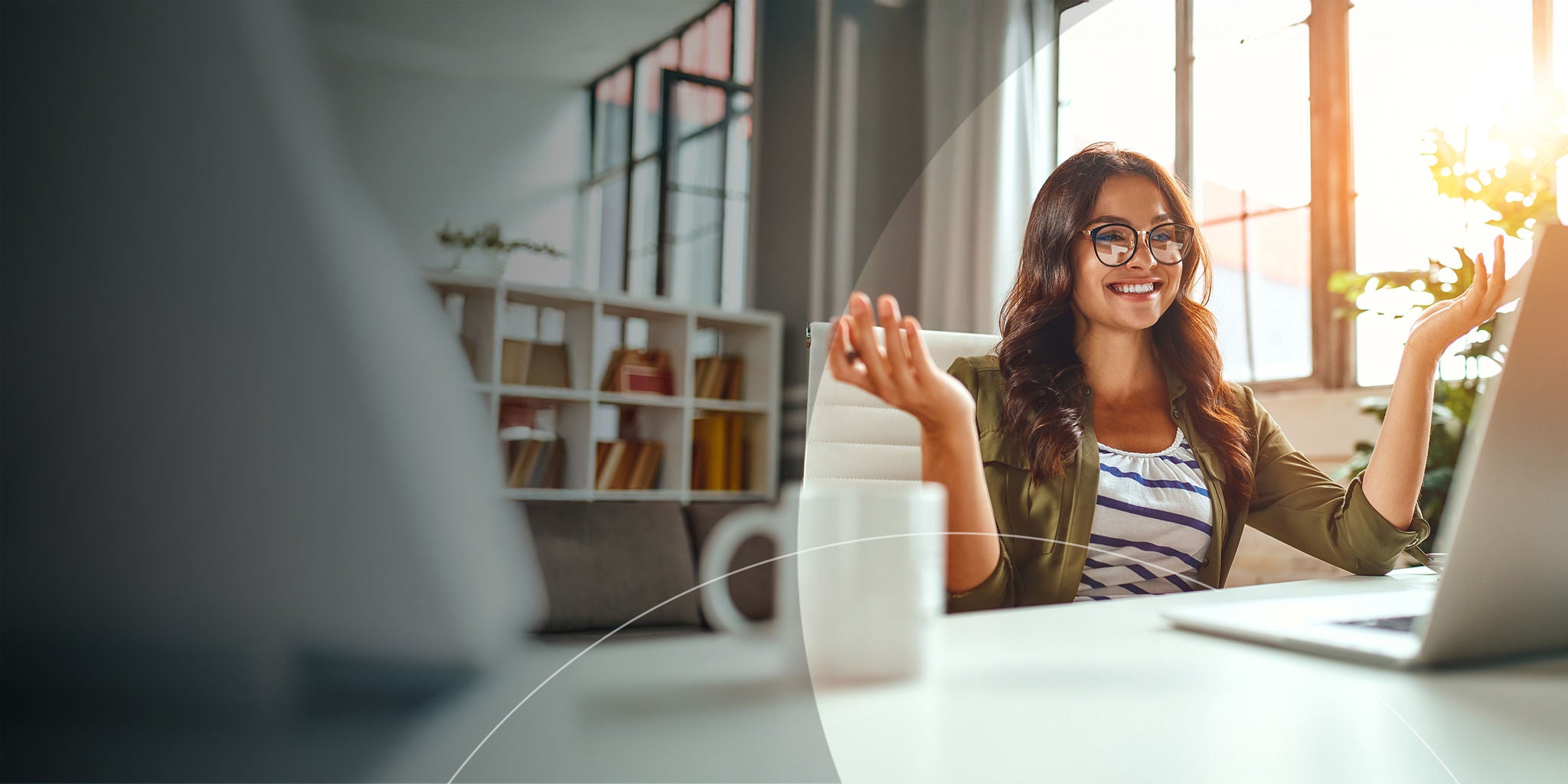 A woman sitting at a desk with her hands raised, looking at her laptop, expressing excitement or surprise.