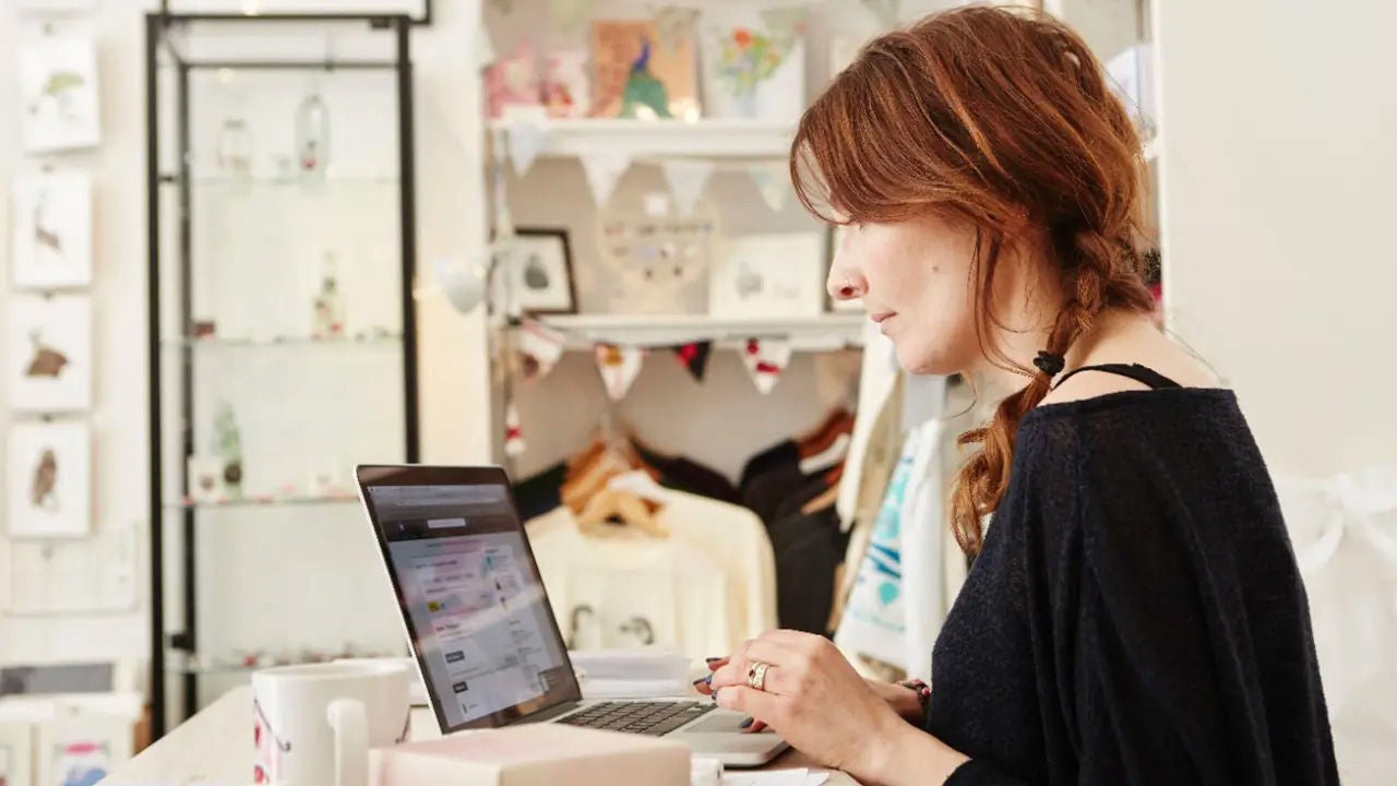 Person working inside small retail shop typing on laptop computer amidst various merchandise displays including clothing items hung up behind them.