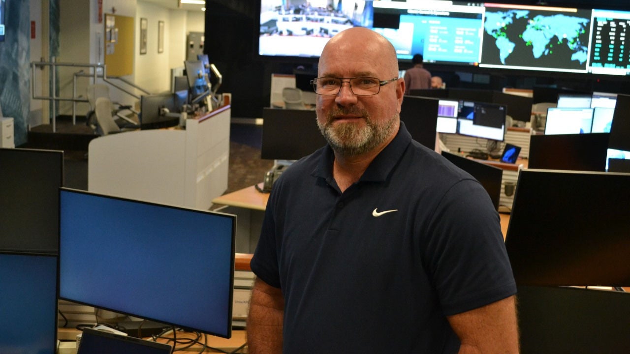 Ken Whitfield stands in front of computers and wall-mounted screens. 