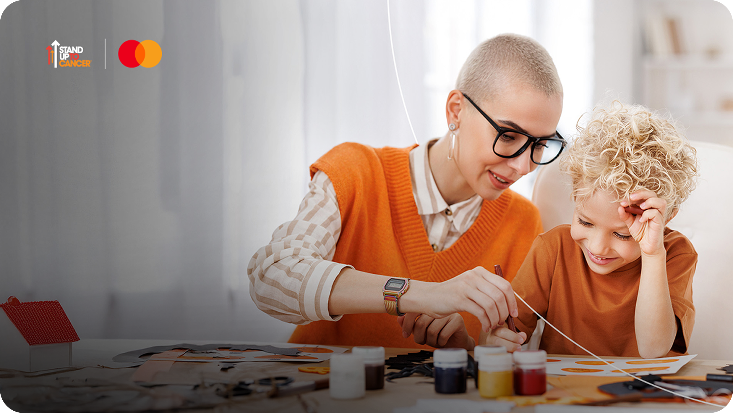 A smiling adult and a young child enjoy a moment together painting, with the Stand Up To Cancer and Mastercard logos visible.