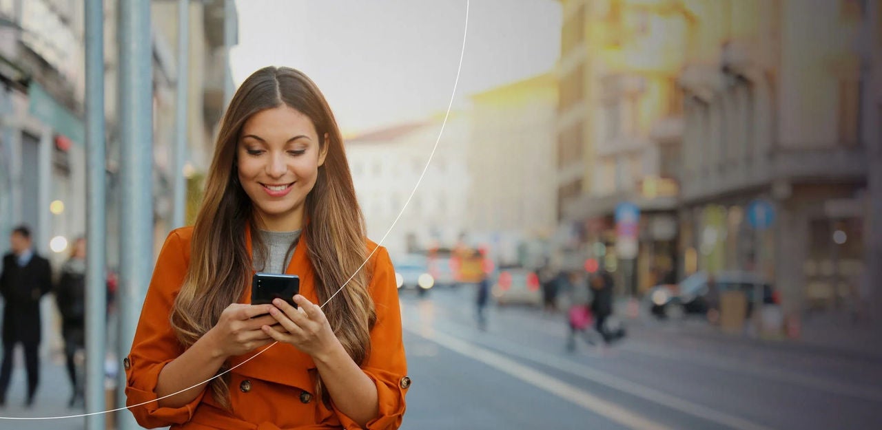 אישה במעיל כתום עומדת ברחוב עירוני, מביטה בטלפון חכם, על רקע עירוני סואן.
