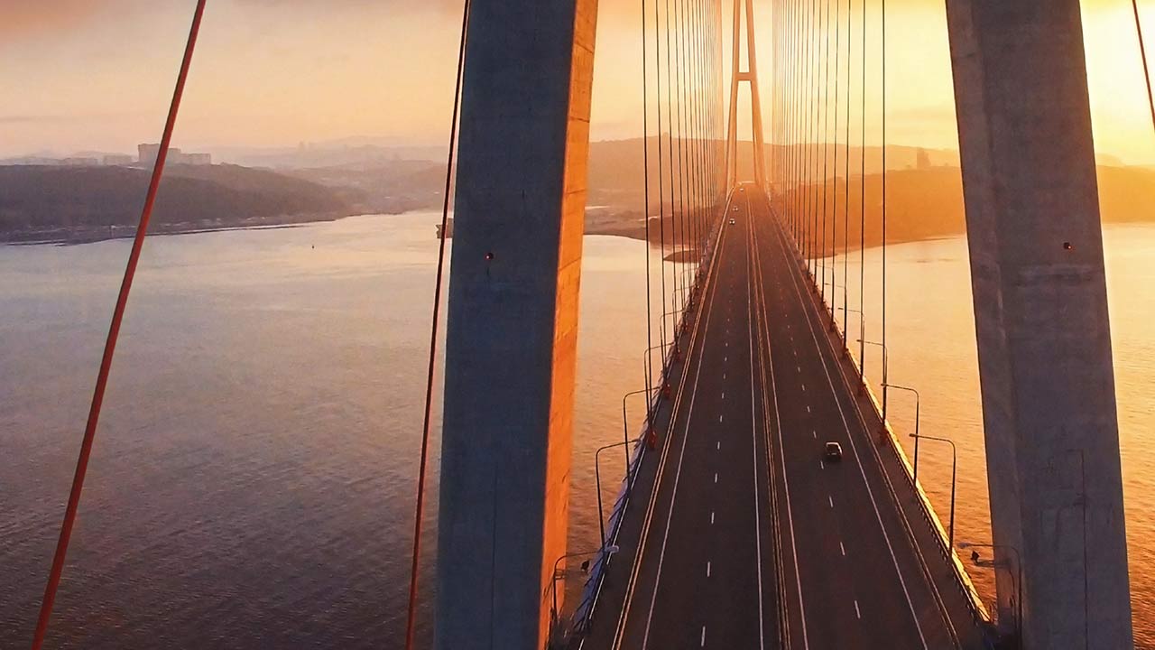 Függőhíd naplementében, felülnézetből, alattuk széles folyóval, a hídon haladó autókkal és aranyszínű fényekkel.