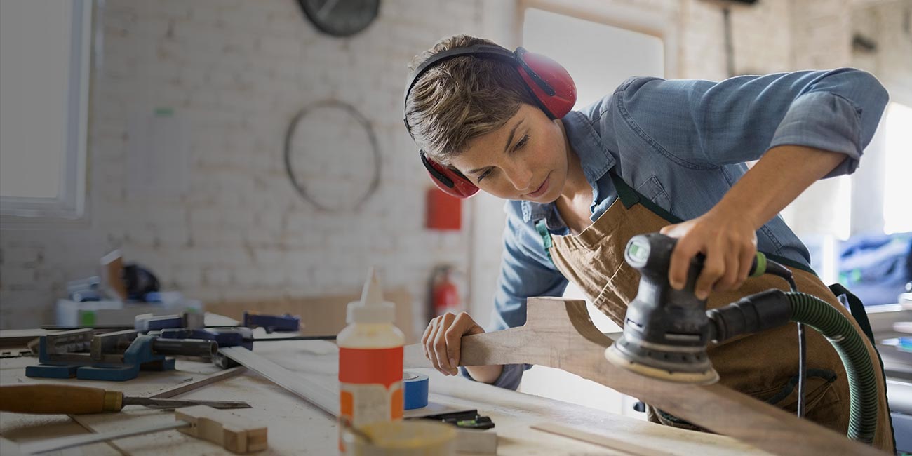 Faipari műhelyben dolgozó nő csiszológéppel simít egy fadarabot, védőfülhallgatót viselve a munkapadnál.