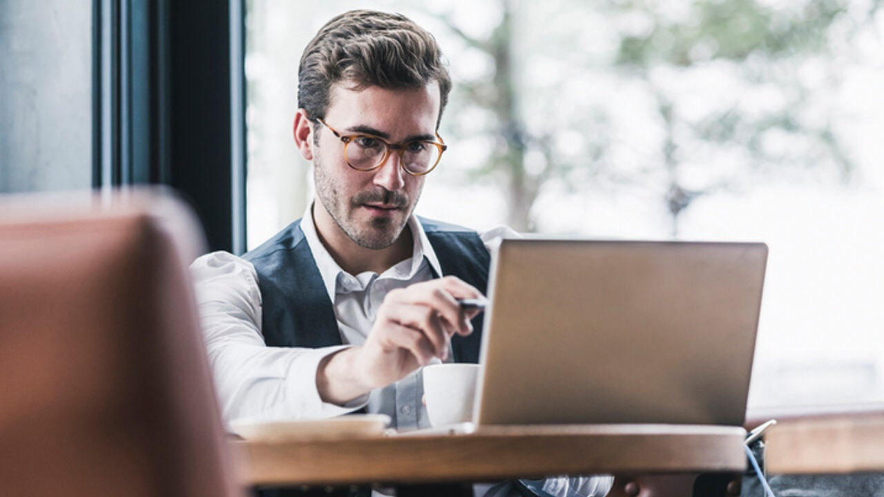 Egy szemüveges férfi kávézóban ülve laptopon dolgozik, egyik kezében tollat tart, az asztalon kávéscsésze látható.