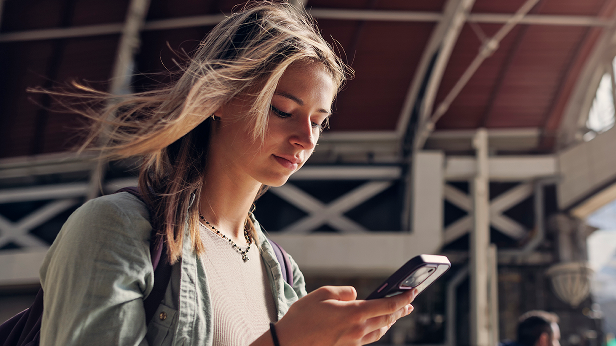 Close-up profile of a young woman with windblown blonde hair looking down at her mobile phone while standing in a busy transit station.