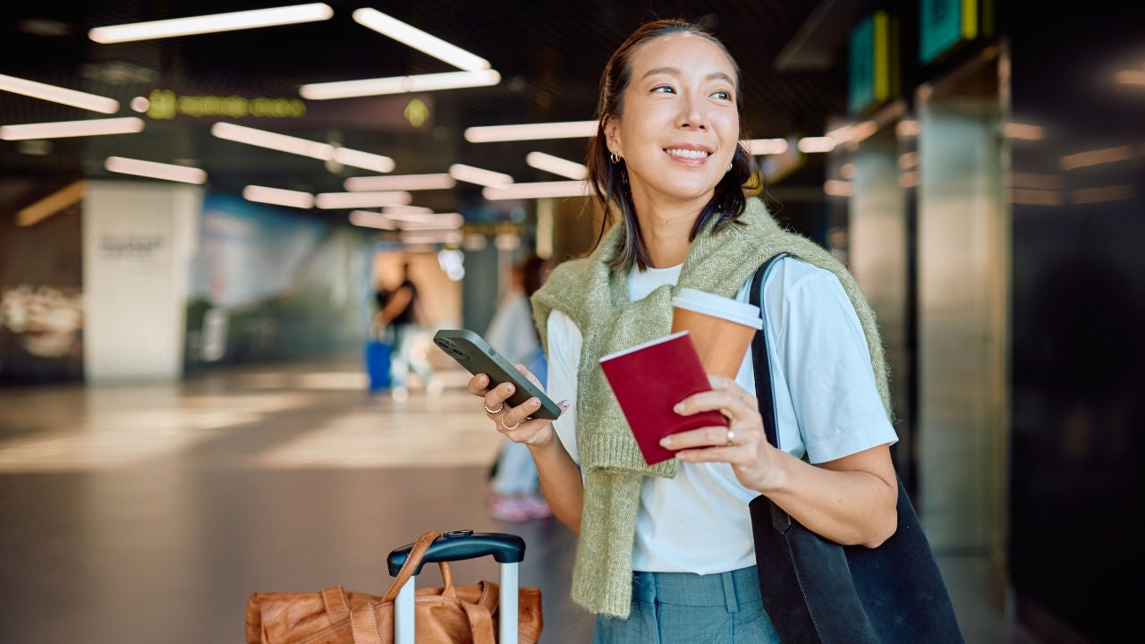 A woman holds a smartphone, passport and coffee cup in her hand with a suitcase next to her in a transit hub. 