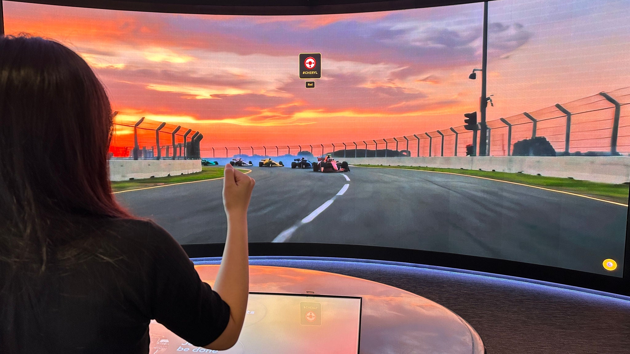A woman raises her fist in front of a large curved screen showing race cars on a track at sunset, with a touchscreen console in front of her.